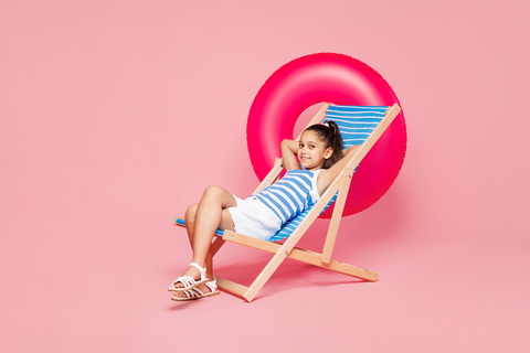 Mädchen in Sommerkleidung auf Liegestuhl mit pinkem Schwimmring im Hintergrund