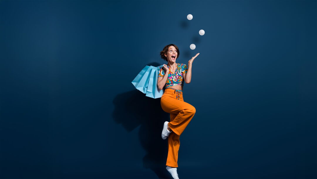 Fröhliche Frau mit Einkaufstaschen springt hoch und jongliert dabei drei Bälle